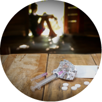 Person in background sitting against doorframe, pills and syringe laying on table in foreground.